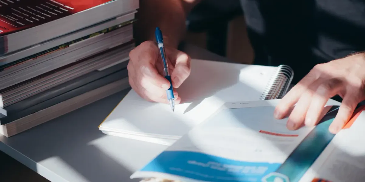 Ręce piszące w notatniku obok stosów książek