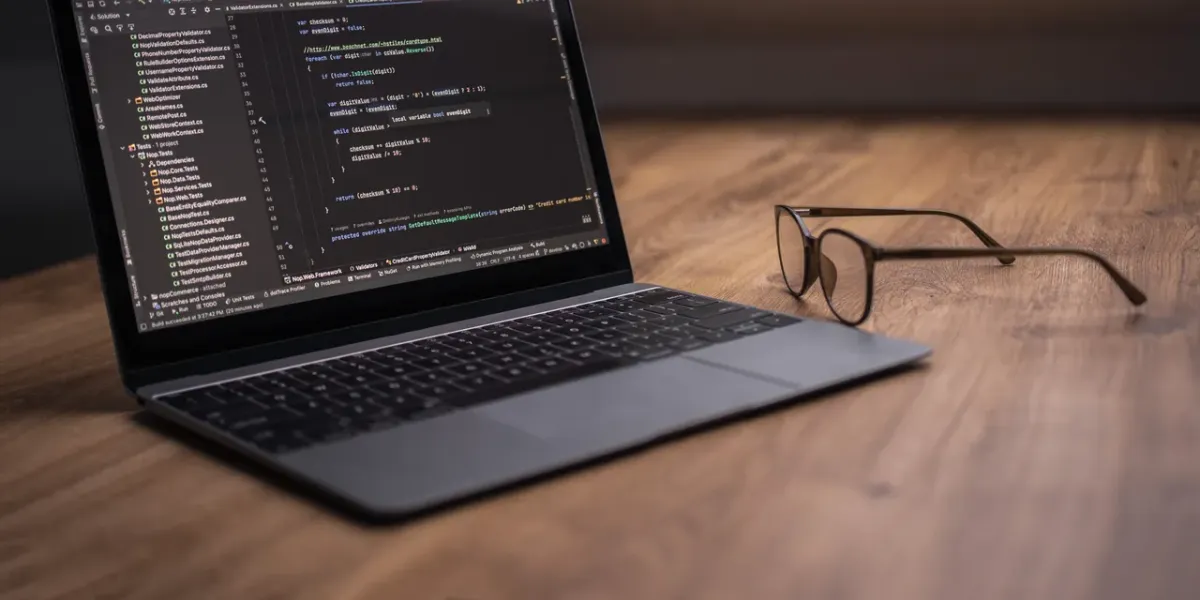 Laptop z otwartym edytorem kodu na drewnianym biurku, obok leżą okulary