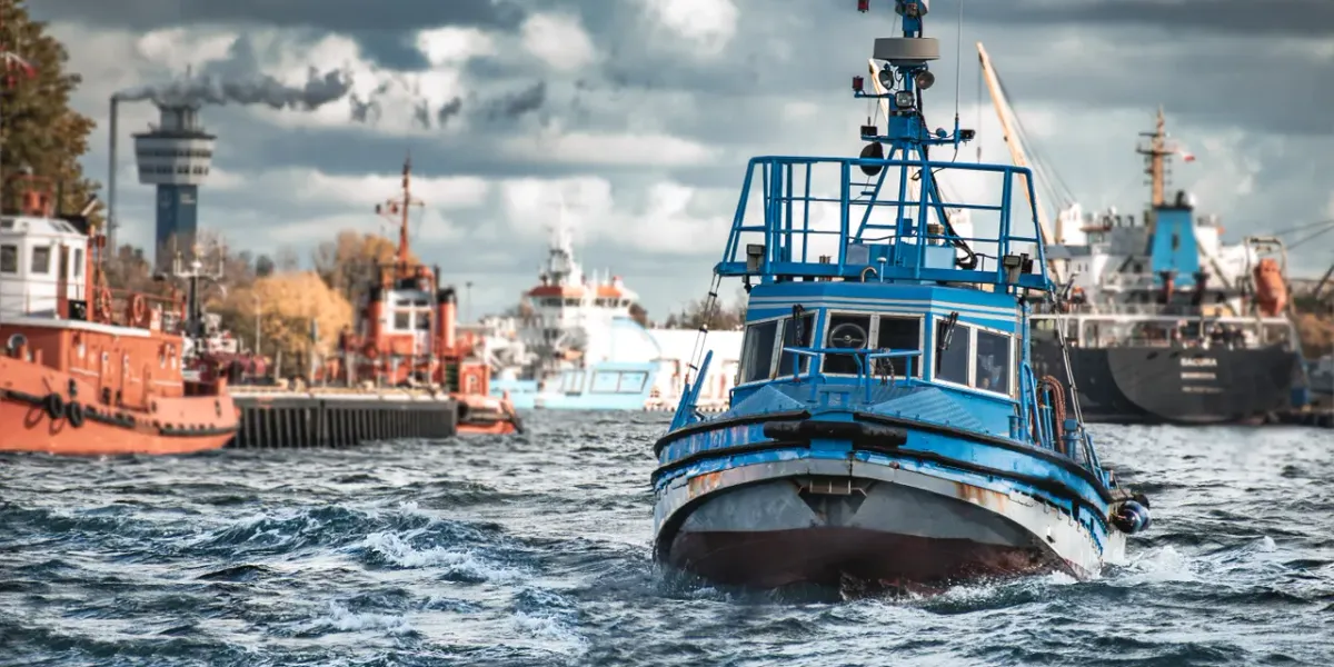 Niebieski holownik płynący po wodach portu, w tle statki i dźwigi