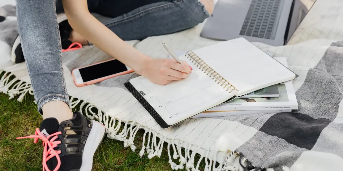 Studentka siedząca na kocu w parku, robi notatki przy laptopie